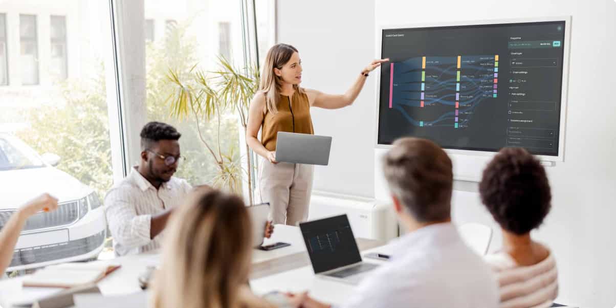 A businesswoman holds a laptop, presenting amoungst a group of colleagues.